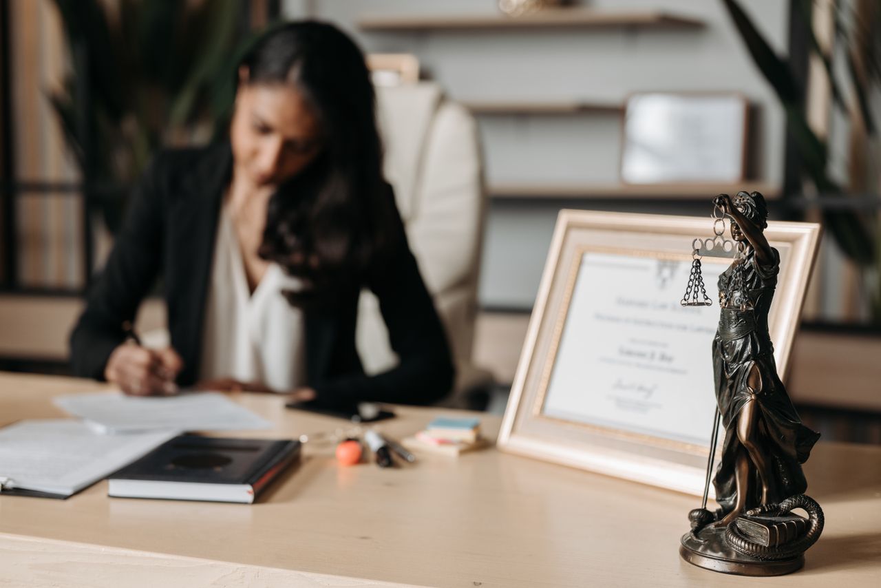 creative Focused woman writing at desk with Lady Justice figurine and certificate in office.
